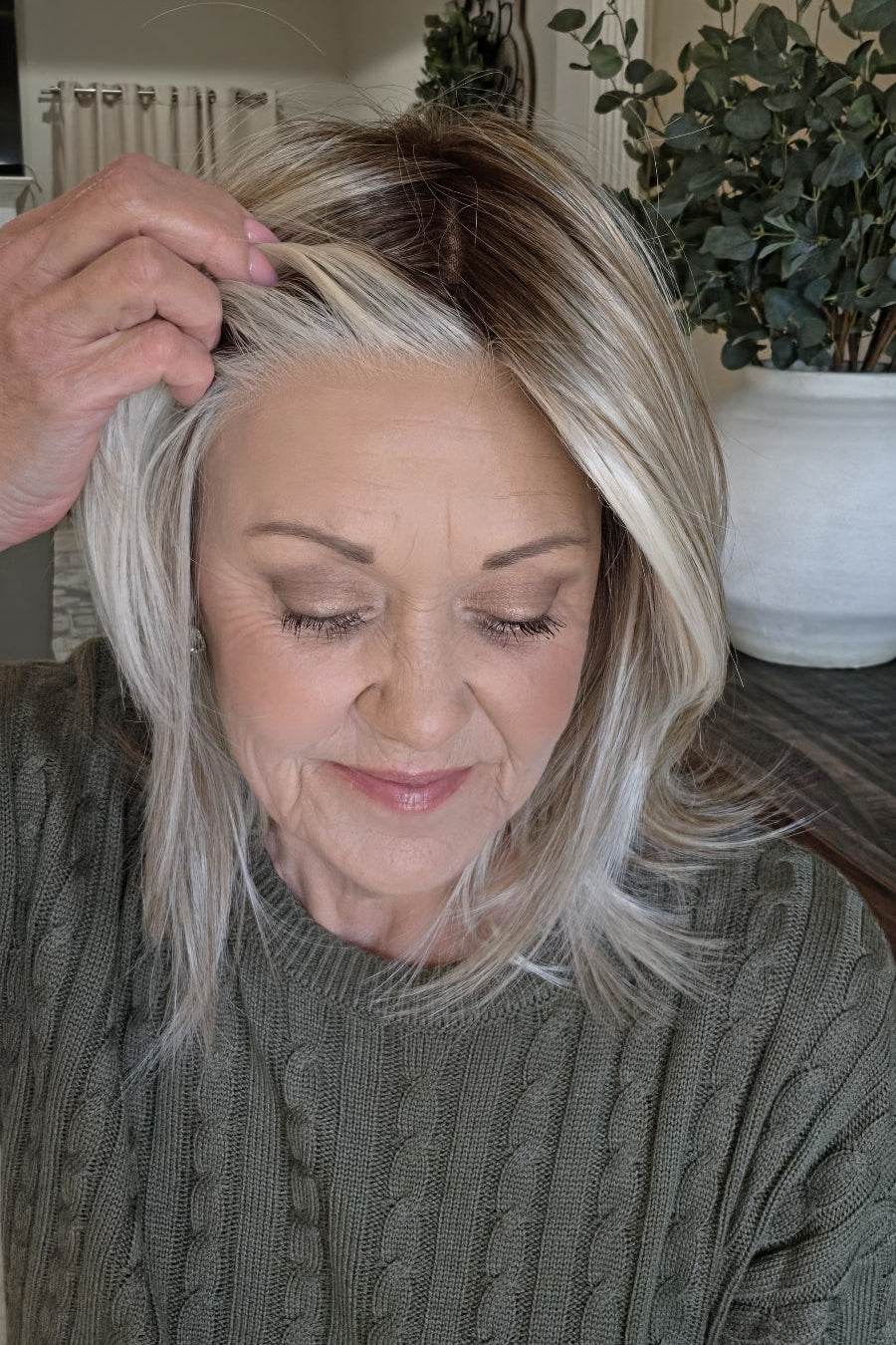 A smiling woman with shoulder-length layered blonde hair wearing an olive green cable-knit sweater (Sloane by Jon Renau in Catalina Blonde).