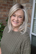 A smiling woman with shoulder-length layered blonde hair wearing an olive green cable-knit sweater (Sloane by Jon Renau in Catalina Blonde).