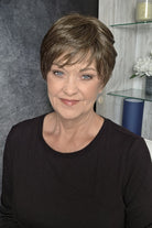 A woman with a short, layered brown and blonde bob hairstyle wearing a black long-sleeve top (Tess by Jon Renau in color Milkshake).