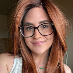 Mackenzie wearing a long, layered auburn wig and black-rimmed glasses, wearing a white knit sleeveless top and gold necklaces, photographed in a casual indoor setting.