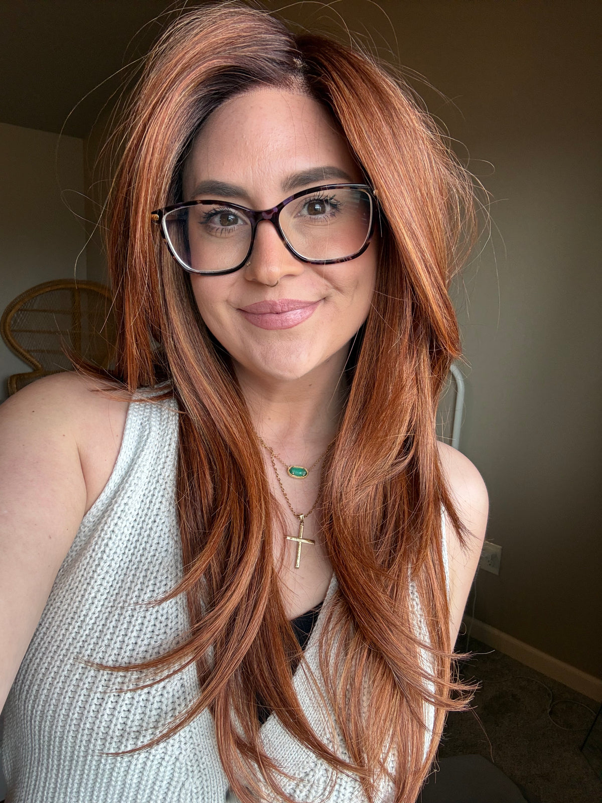 A woman with long, wavy coppery auburn hair and black-rimmed glasses, wearing a white knit sleeveless top. She is photographed in a casual indoor setting.