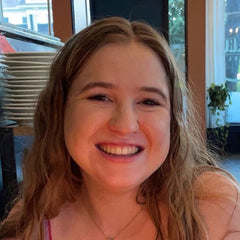 Megan with long light brown hair sits at a table in a restaurant or cafe, wearing a pink and purple patterned spaghetti print tank top. In front of her is a plate with chocolate-covered pastries dusted with powdered sugar, a small cup of dipping sauce, and a single lit candle.