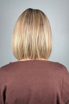 A smiling blonde woman with shoulder-length straight hair and bangs (wearing Miss Comfort by Ellen Wille in color Caramel Rooted). 