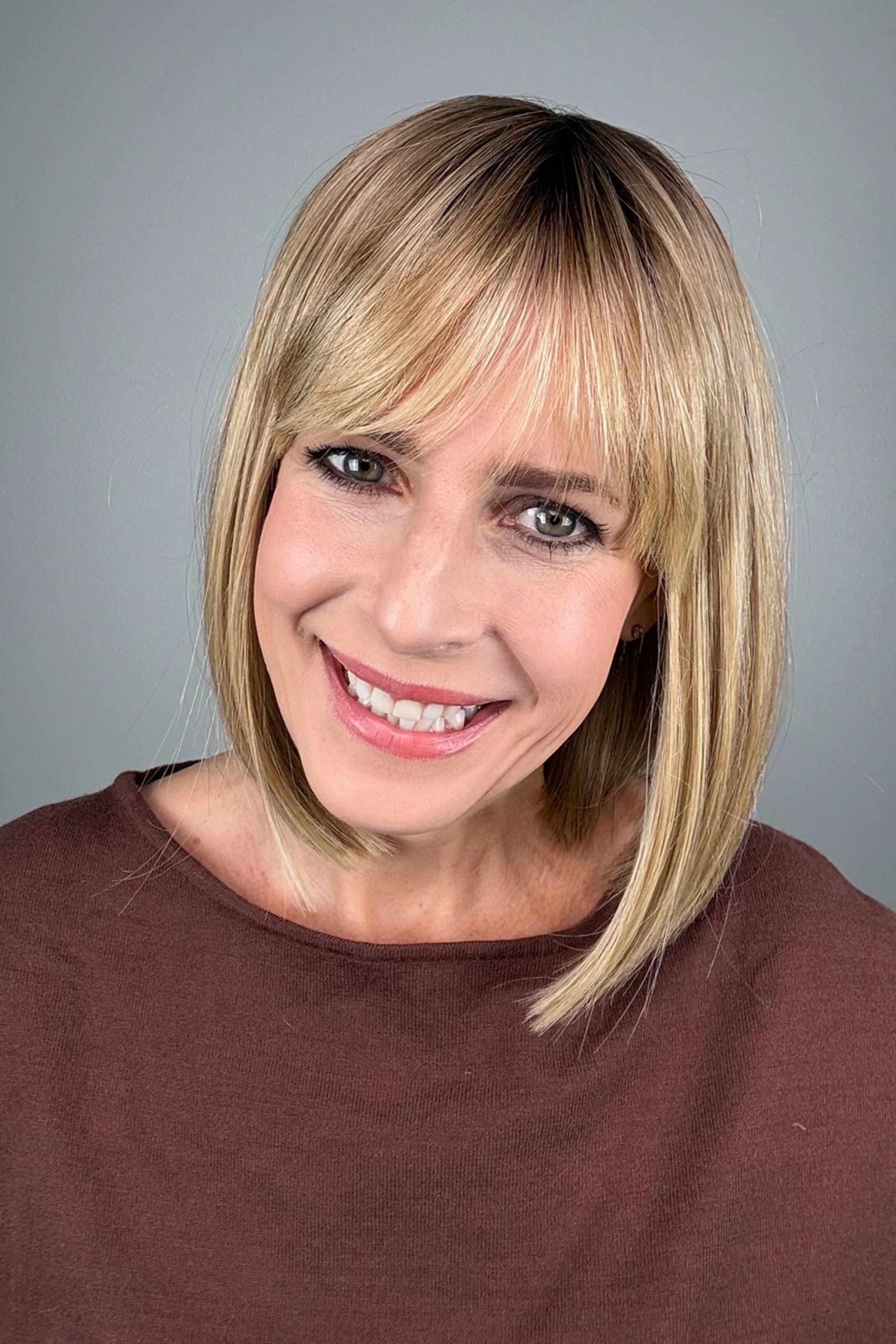 A smiling blonde woman with shoulder-length straight hair and bangs (wearing Miss Comfort by Ellen Wille in color Caramel Rooted). 