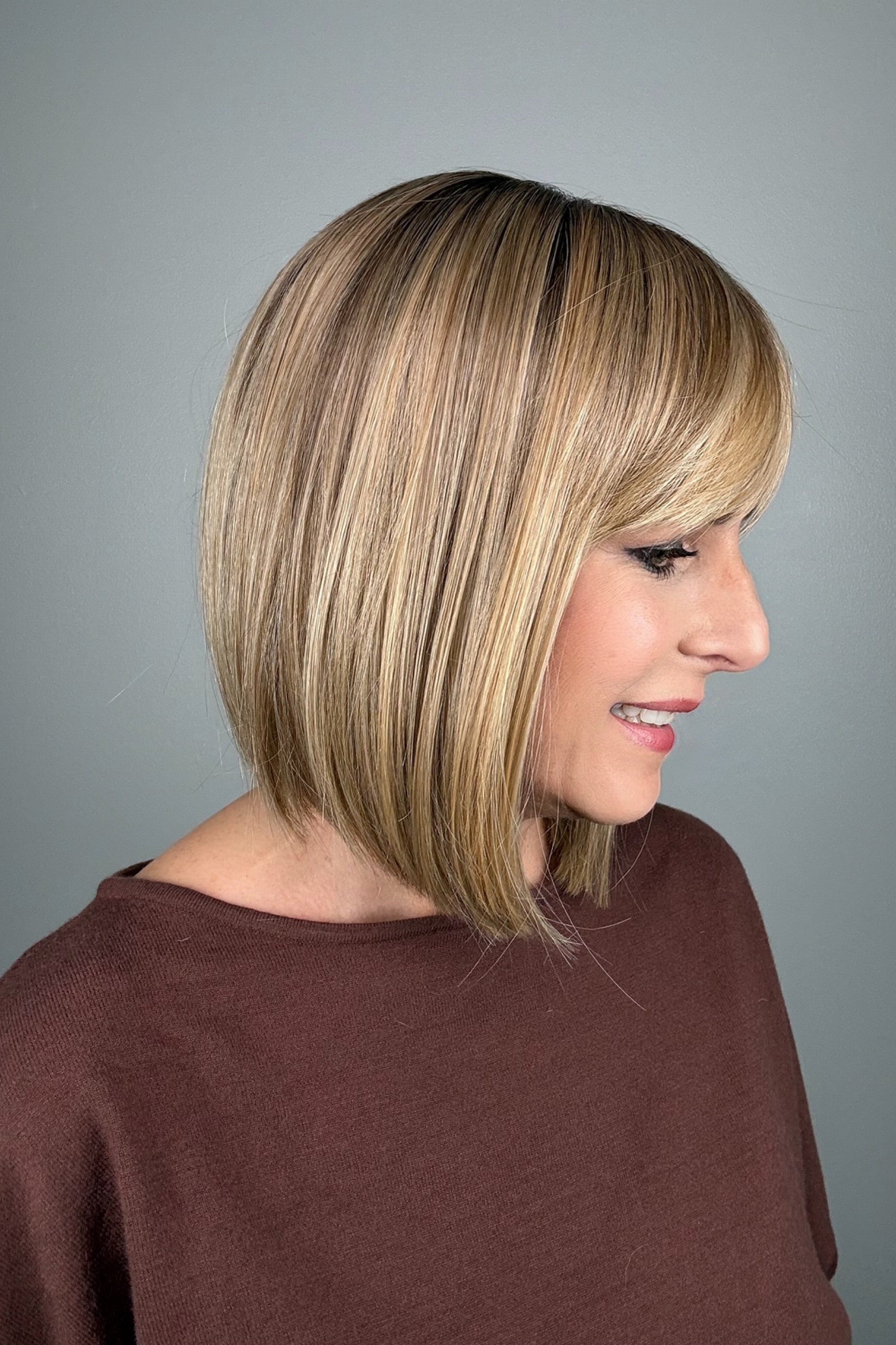 A smiling blonde woman with shoulder-length straight hair and bangs (wearing Miss Comfort by Ellen Wille in color Caramel Rooted). 