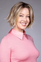 Woman wearing Oslo heat friendly synthetic wig in color raw sugar blonde by Belle Tress with a light grey background and pink shirt