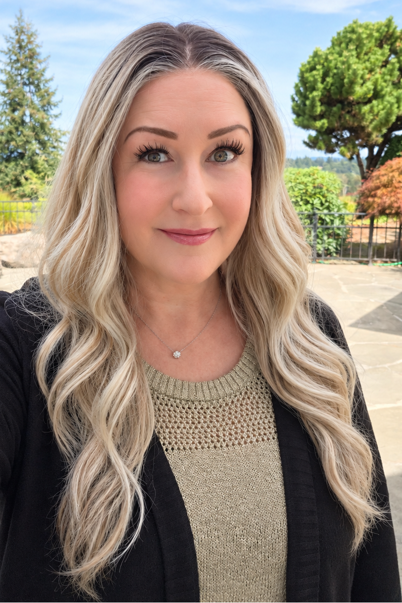 A woman wearing Bradley synthetic wig by PUSH in color Champagne Blonde Rooted is shown in a selfie-style outdoor portrait. She is wearing a black cardigan over a beige knit top, with a backyard pool, trees, and blue sky in the background.
