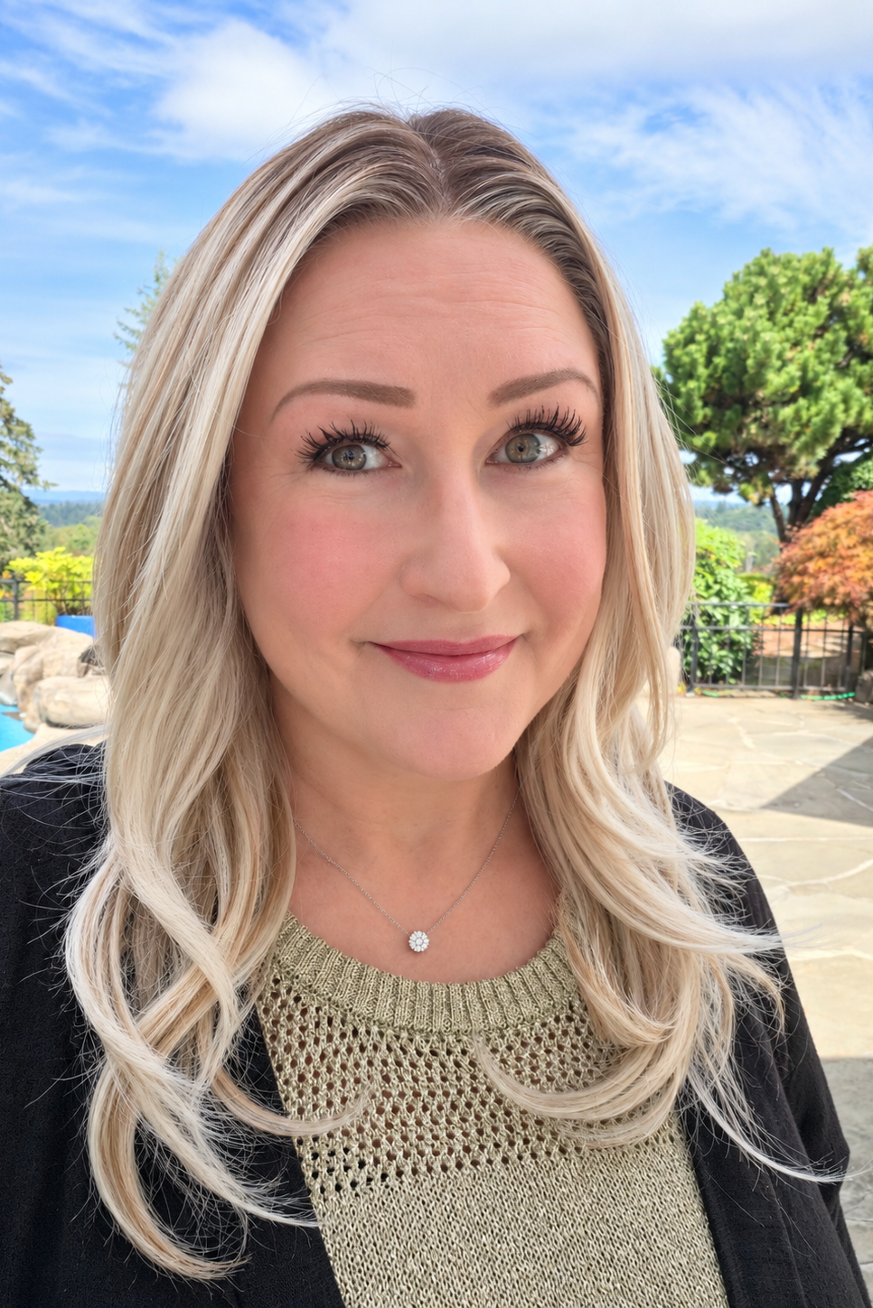 A woman wearing Olivia by PUSH - a long synthetic wig in color Pearl Silk Rooted in a close-up portrait outdoors. She is wearing a black cardigan over a textured beige knit top and a small silver necklace.