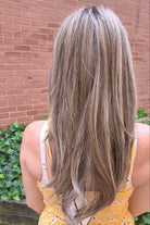 A woman with long highlighted brown and blonde wig wearing a yellow sleeveless top with black polka dots and brown buttons, photographed outdoors against a brick wall and green ivy. Scene Stealer by Raquel Welch in Shaded Cappuccino.