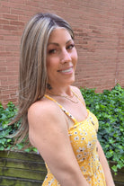 A woman with long highlighted brown and blonde wig wearing a yellow sleeveless top with black polka dots and brown buttons, photographed outdoors against a brick wall and green ivy. Scene Stealer by Raquel Welch in Shaded Cappuccino.