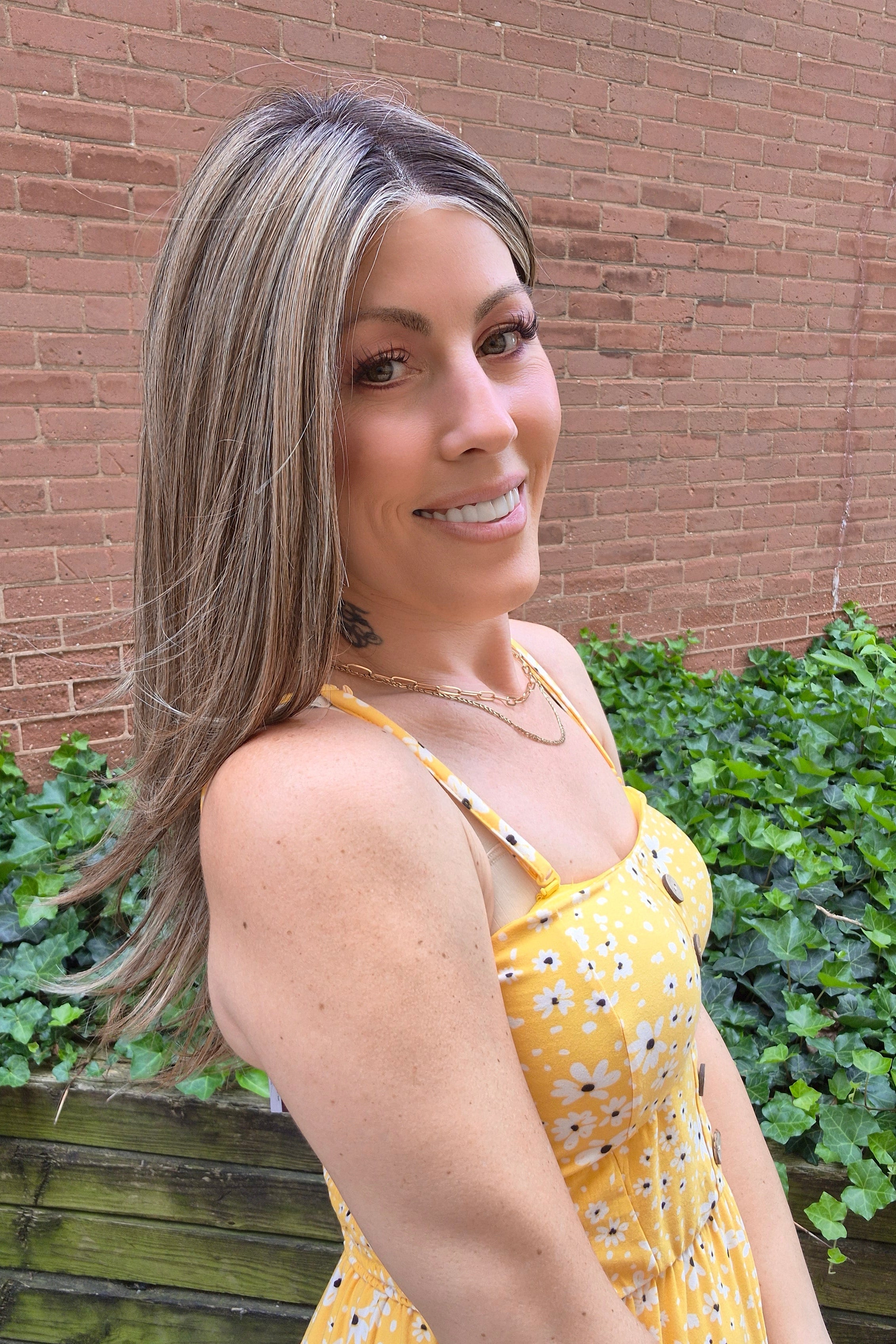 A woman with long highlighted brown and blonde wig wearing a yellow sleeveless top with black polka dots and brown buttons, photographed outdoors against a brick wall and green ivy. Scene Stealer by Raquel Welch in Shaded Cappuccino.