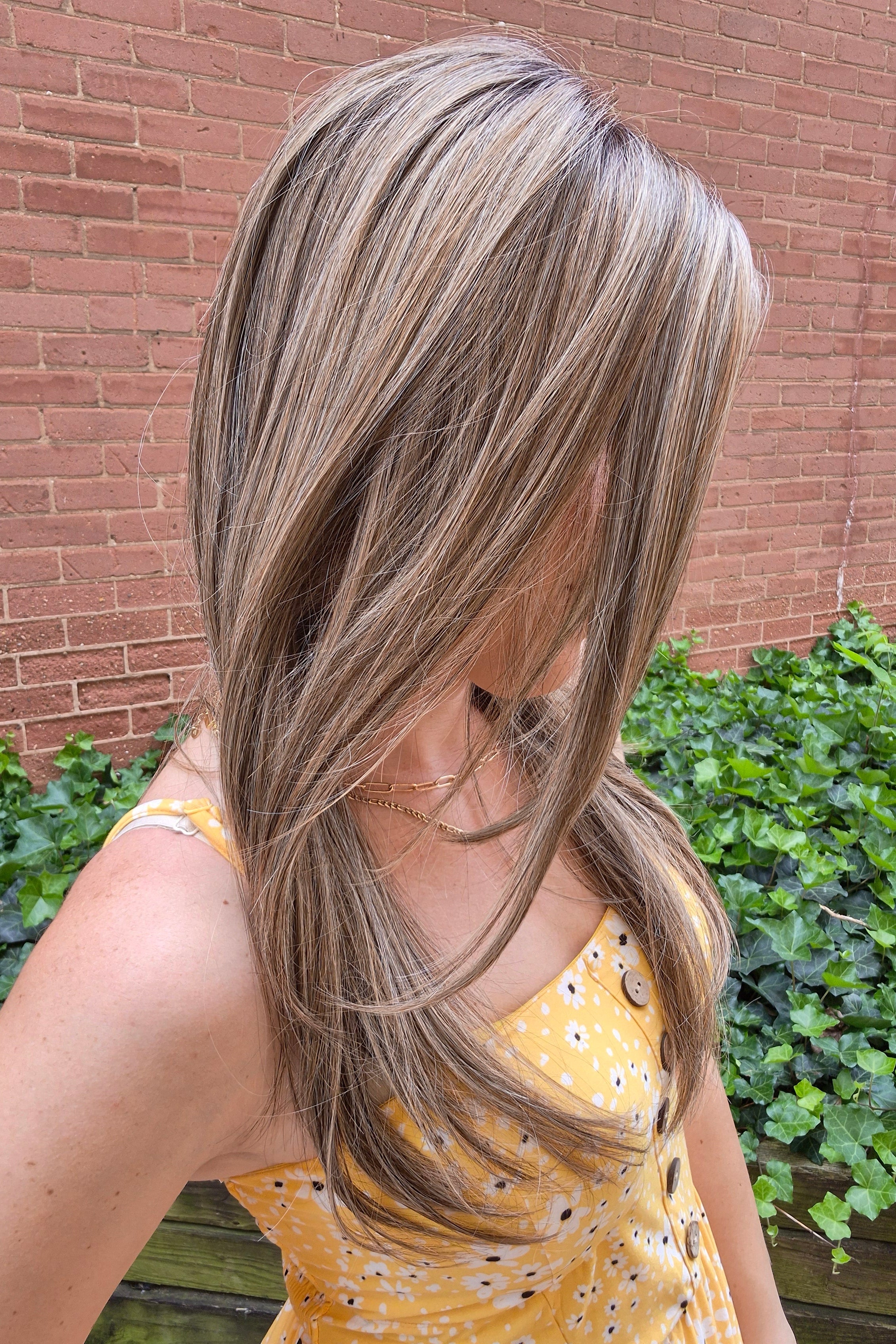 A woman with long highlighted brown and blonde wig wearing a yellow sleeveless top with black polka dots and brown buttons, photographed outdoors against a brick wall and green ivy. Scene Stealer by Raquel Welch in Shaded Cappuccino.