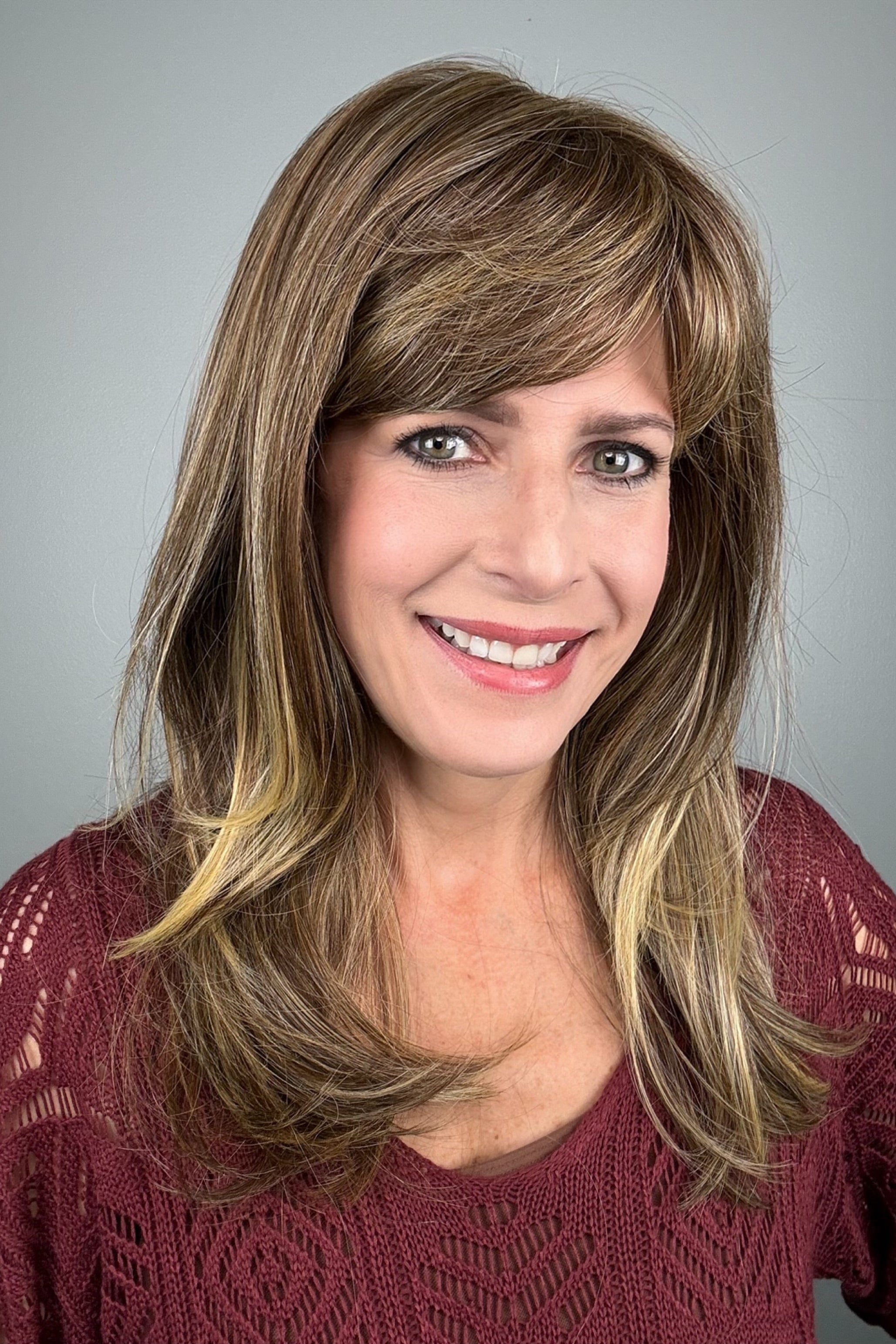 A woman with shoulder-length layered brown hair with blonde highlights and straight-cut bangs (wearing Revelry by Gabor in color Brown Blonde).