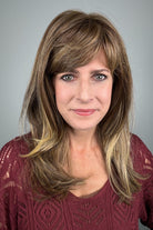 A woman with shoulder-length layered brown hair with blonde highlights and straight-cut bangs (wearing Revelry by Gabor in color Brown Blonde).