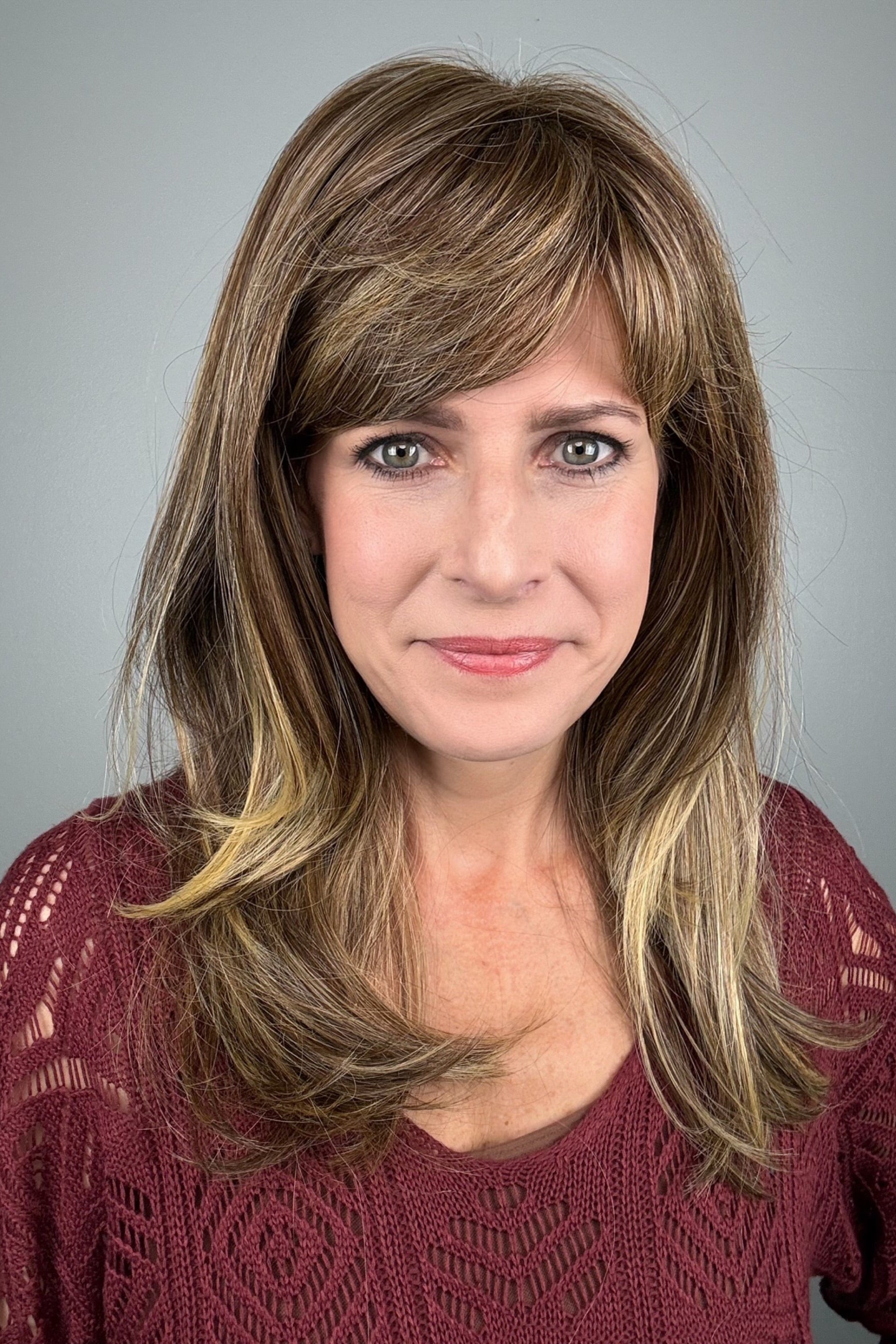A woman with shoulder-length layered brown hair with blonde highlights and straight-cut bangs (wearing Revelry by Gabor in color Brown Blonde).