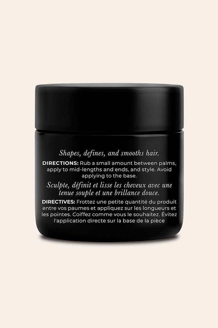 A black cosmetic jar with a matte black lid, featuring white text detailing directions for hair shaping and definition, set against a solid light beige background.