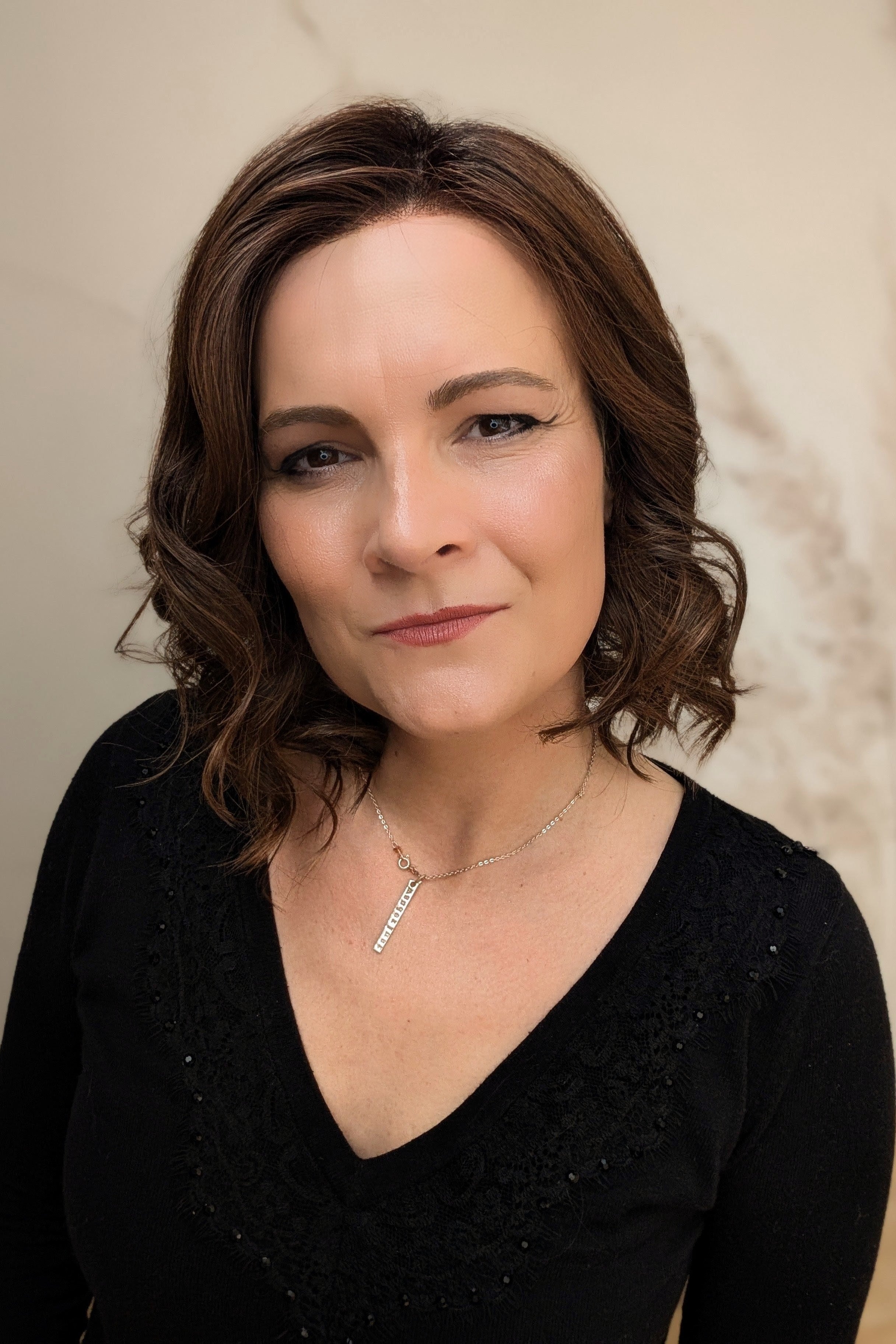 A woman with shoulder-length brown curly hair wearing a black long-sleeve top (Streep by Belle Tress in color Soft Amber Brown).