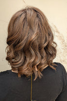 A woman with shoulder-length brown curly hair wearing a black long-sleeve top (Streep by Belle Tress in color Soft Amber Brown).