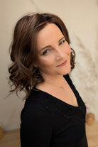 A woman with shoulder-length brown curly hair wearing a black long-sleeve top (Streep by Belle Tress in color Soft Amber Brown).