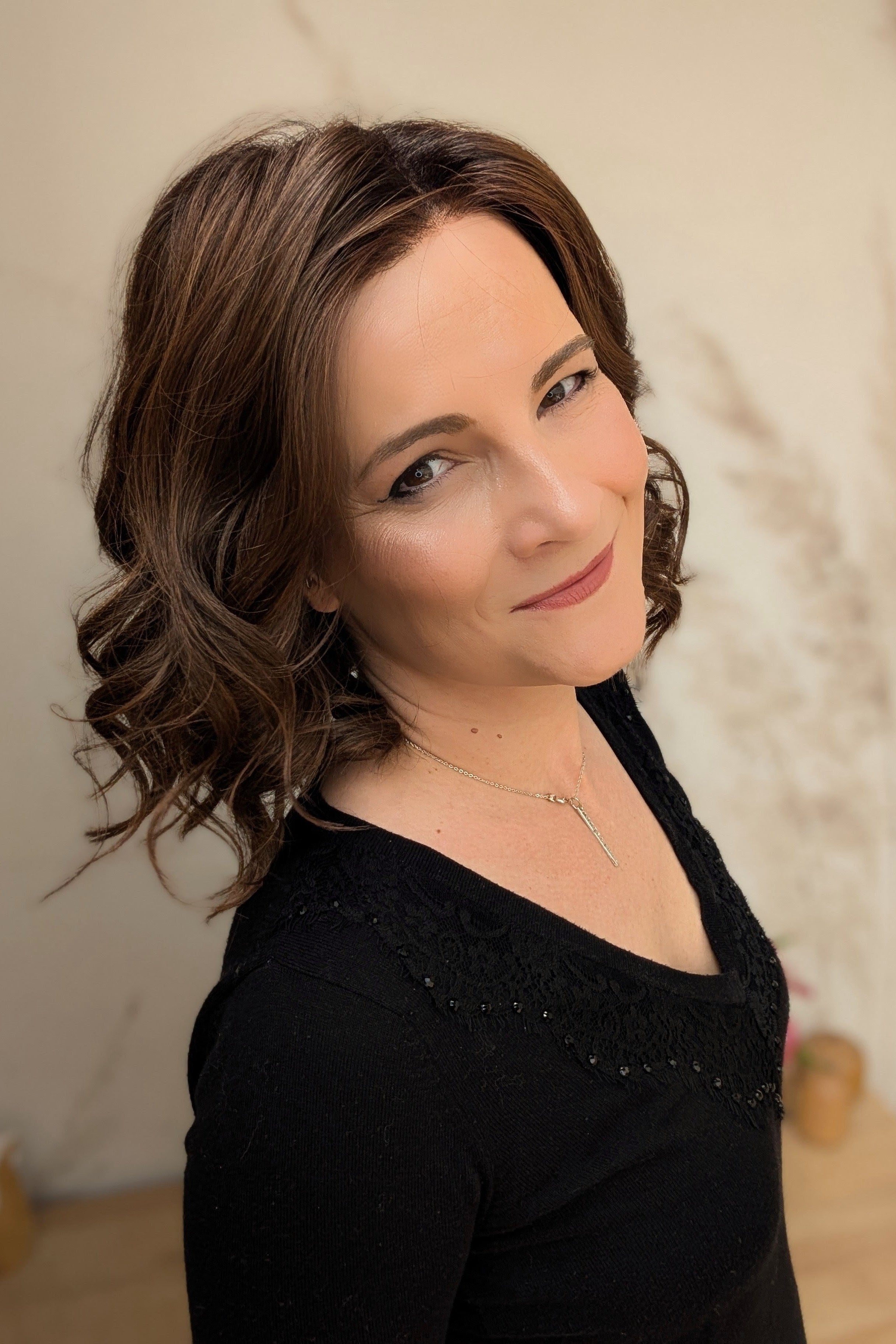 A woman with shoulder-length brown curly hair wearing a black long-sleeve top (Streep by Belle Tress in color Soft Amber Brown).