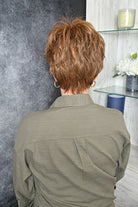A woman with short, layered auburn hair wearing an olive green button-up shirt over a white top (Wearing Kris Wig by Tony of Beverly in color Wineberry).