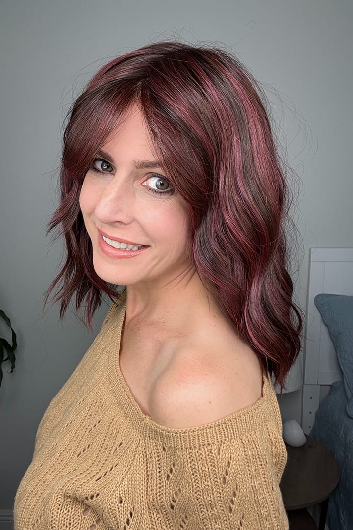 Woman wearing shoulder-length wavy wig with purple highlights (Smooth Wavez by Rene of Paris in color Plum Brown R)