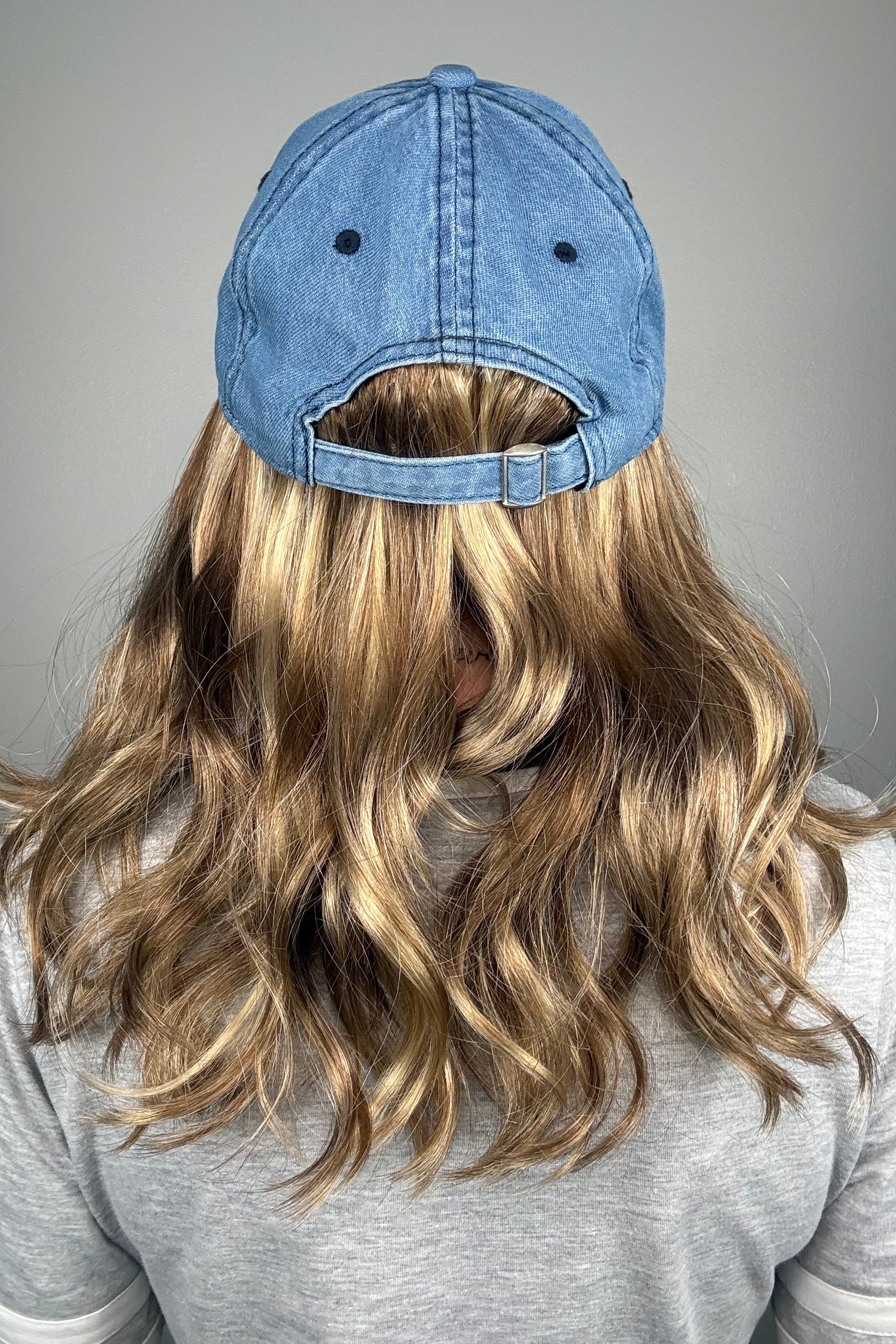 A woman with dark blonde hair wearing a blue denim baseball cap and a gray V-neck top (wearing Wavy Hat Denim in color 12/26H).