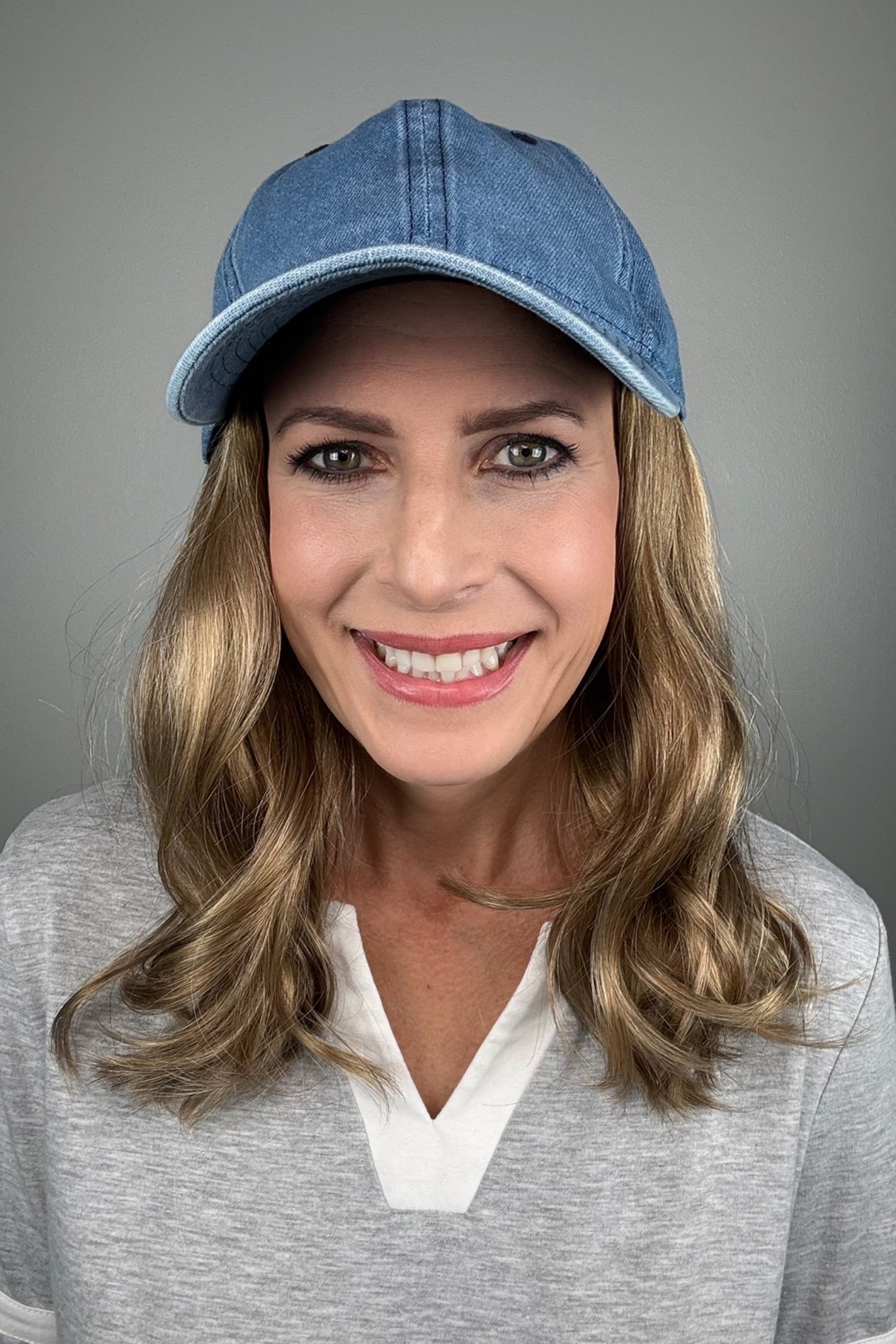 A woman with dark blonde hair wearing a blue denim baseball cap and a gray V-neck top (wearing Wavy Hat Denim in color 12/26H).