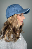 A woman with dark blonde hair wearing a blue denim baseball cap and a gray V-neck top (wearing Wavy Hat Denim in color 12/26H).