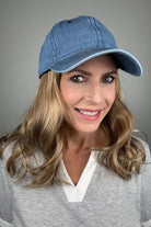 A woman with dark blonde hair wearing a blue denim baseball cap and a gray V-neck top (wearing Wavy Hat Denim in color 12/26H).