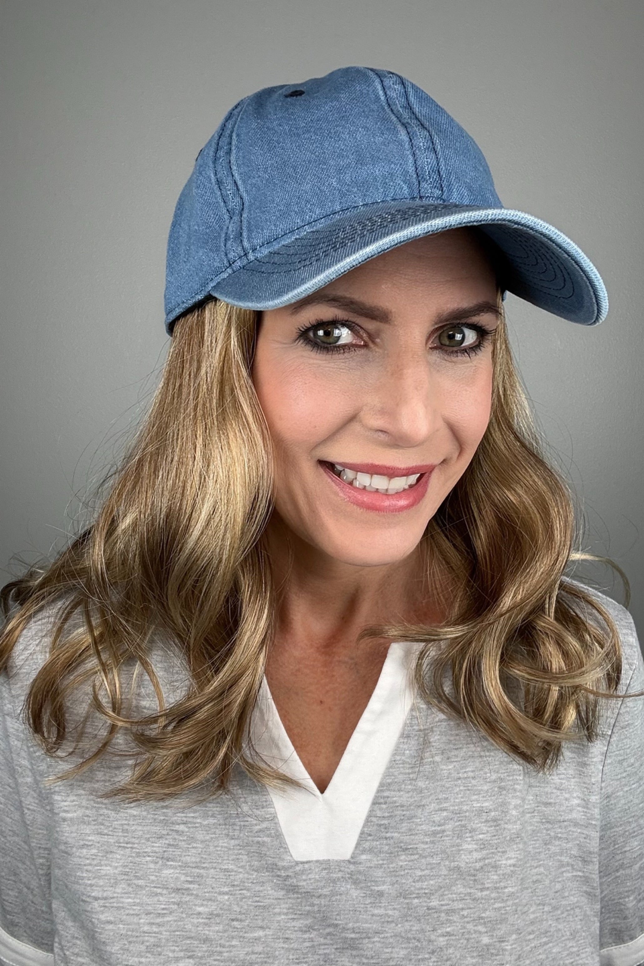 A woman with dark blonde hair wearing a blue denim baseball cap and a gray V-neck top (wearing Wavy Hat Denim in color 12/26H).