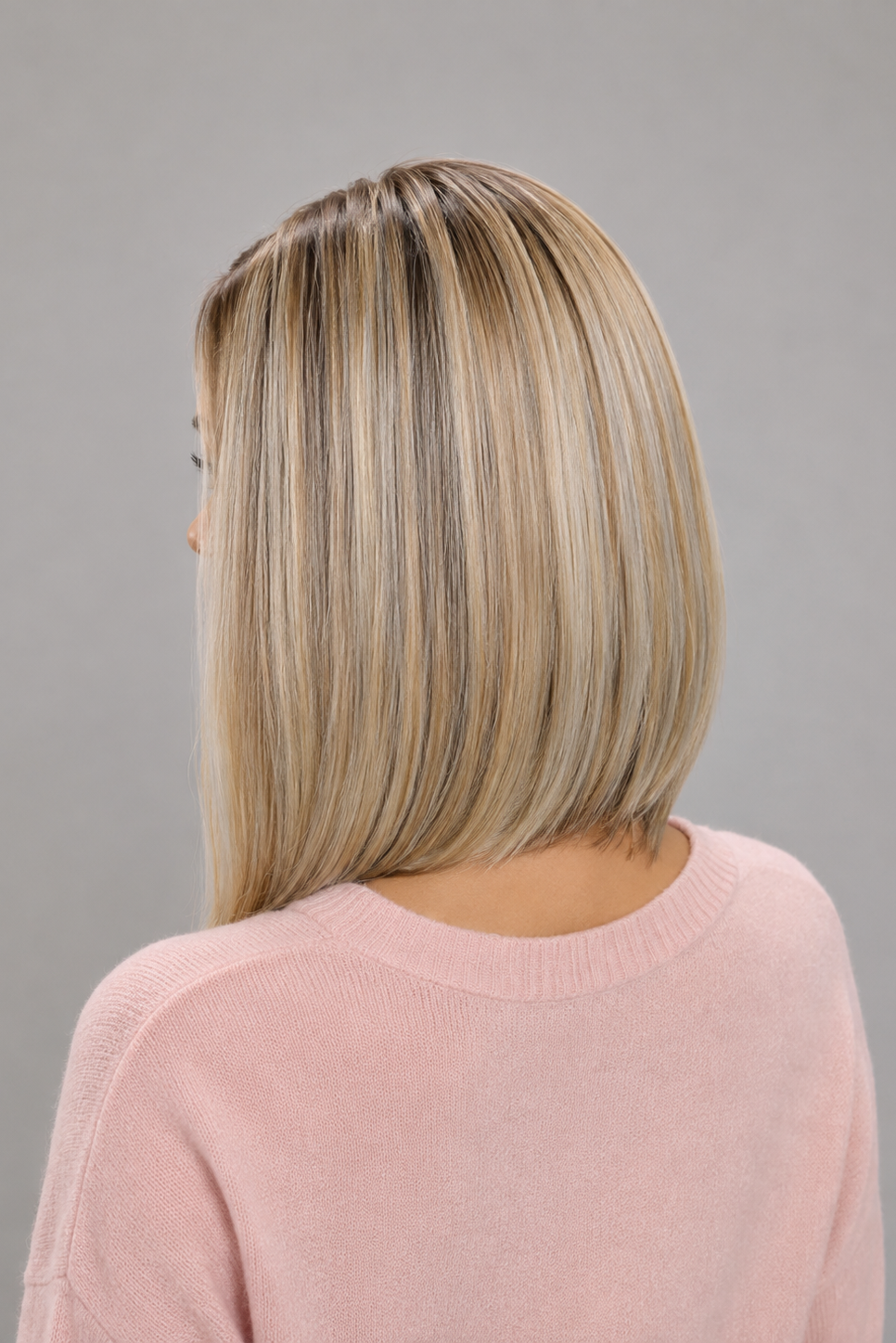 Woman wearing Adelina synthetic wig by PUSH in color white champagne rooted in a light pink v-neck sweater against a light grey background - back view