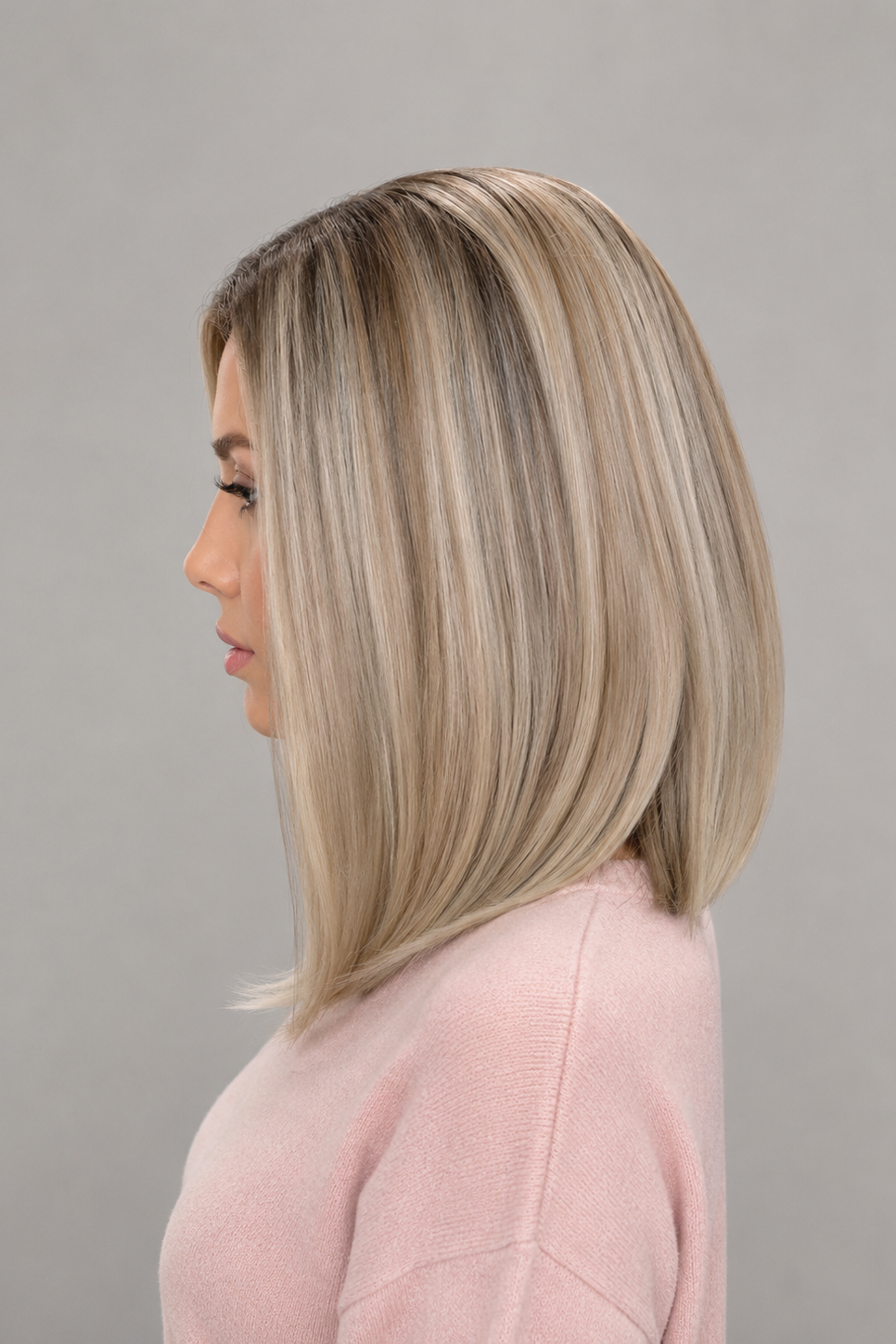 Woman wearing Adelina synthetic wig by PUSH in color white champagne rooted in a light pink v-neck sweater against a light grey background - left side view