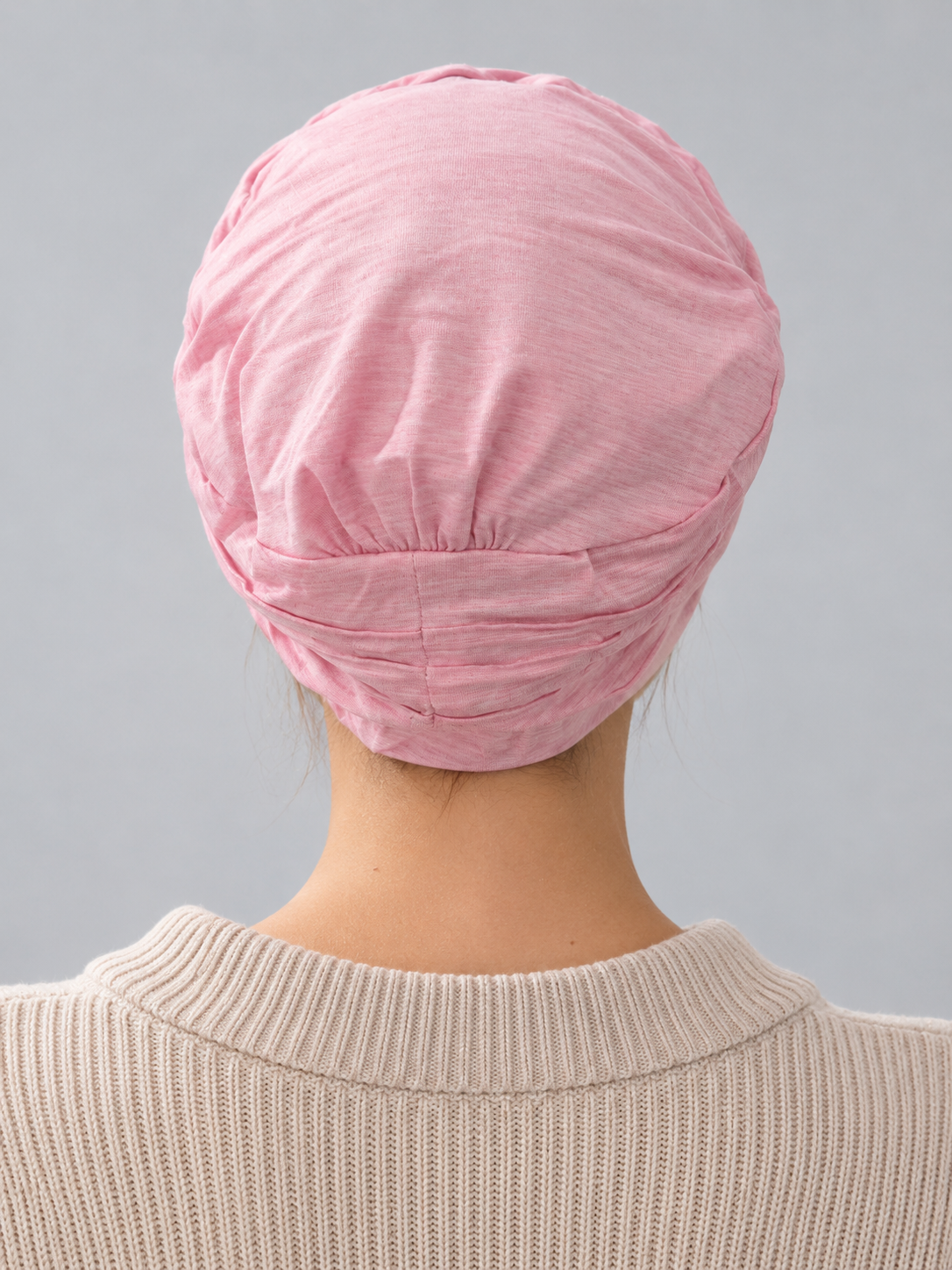 A woman wearing a rose melange turban-style cap, paired with a beige ribbed zip-up sweater, against a solid gray studio background. Back view