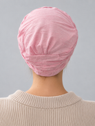 A woman wearing a rose melange turban-style cap, paired with a beige ribbed zip-up sweater, against a solid gray studio background. Back view
