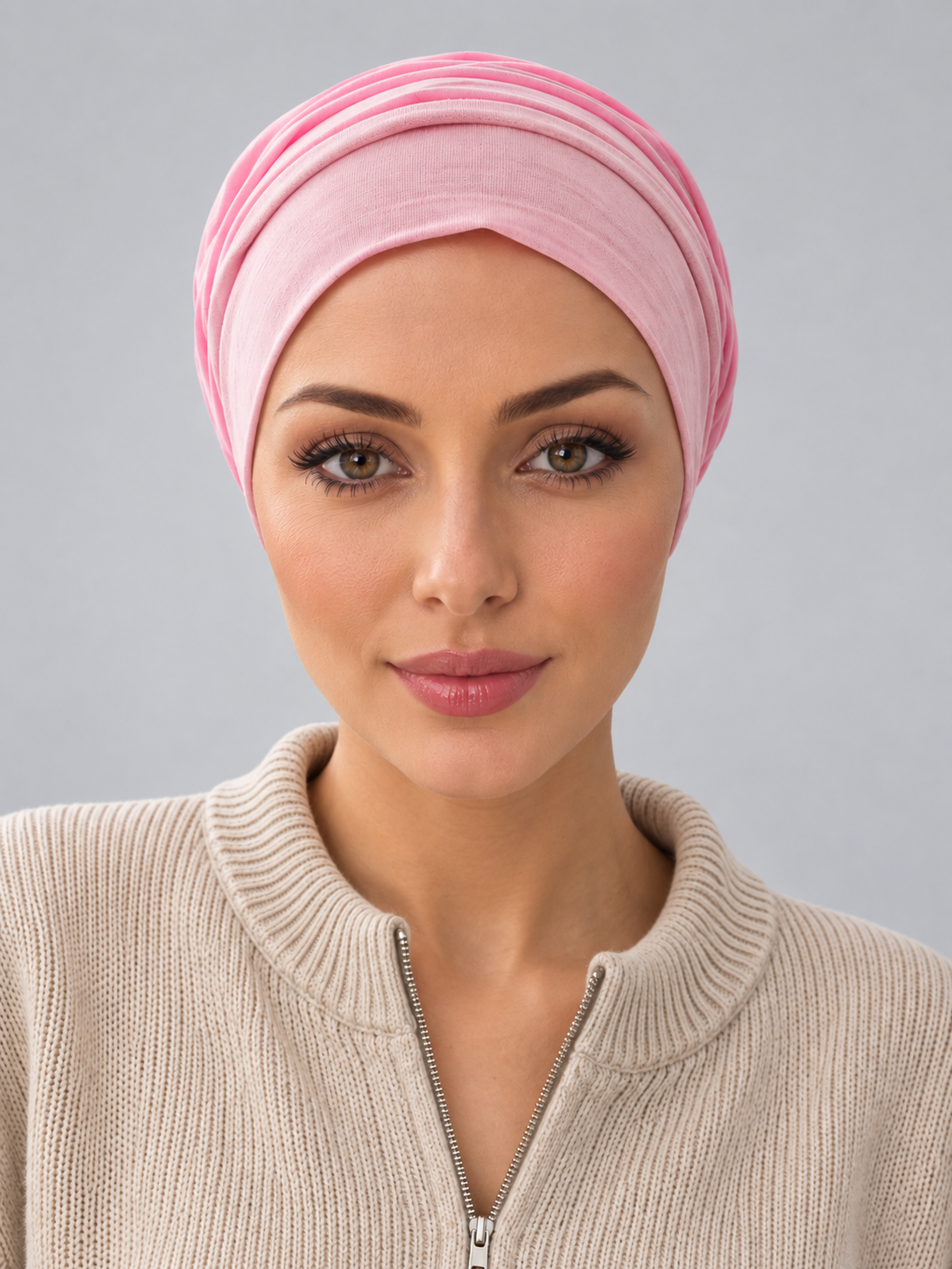 A woman wearing a rose melange turban-style cap, paired with a beige ribbed zip-up sweater, against a solid gray studio background. Front view
