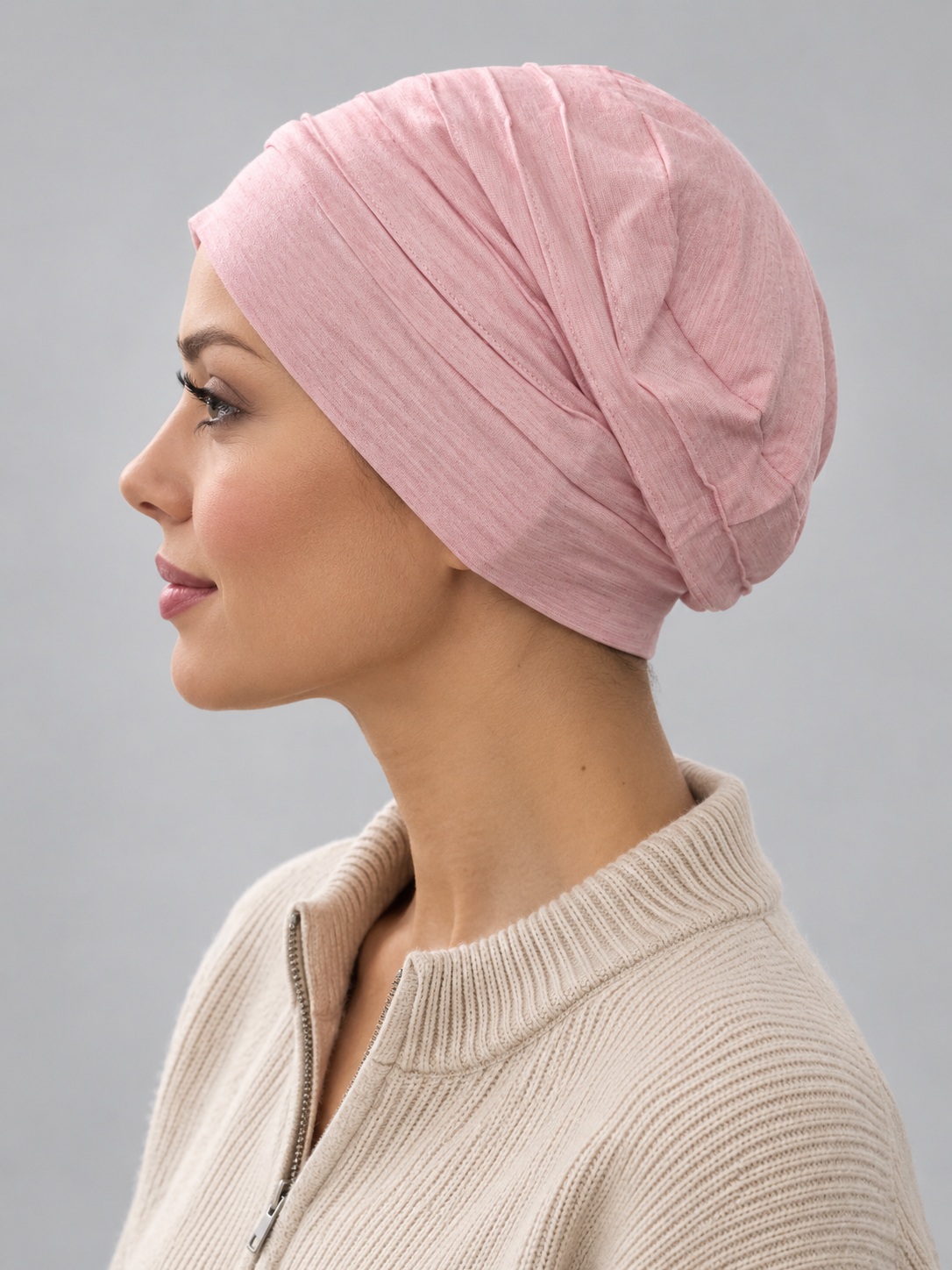 A woman wearing a rose melange turban-style cap, paired with a beige ribbed zip-up sweater, against a solid gray studio background. Left side view.