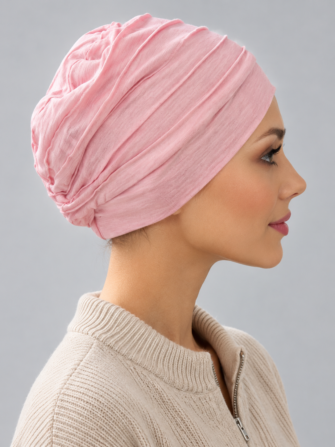 A woman wearing a rose melange turban-style cap, paired with a beige ribbed zip-up sweater, against a solid gray studio background. Right side view