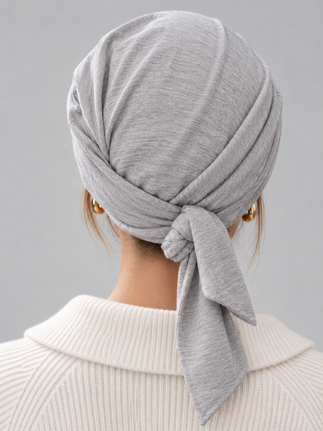 A woman wearing a light gray knitted turban headscarf, paired with a cream ribbed knit top, against a solid gray studio background. Back view