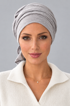 A woman wearing a light gray knitted turban headscarf, paired with a cream ribbed knit top, against a solid gray studio background. Front view