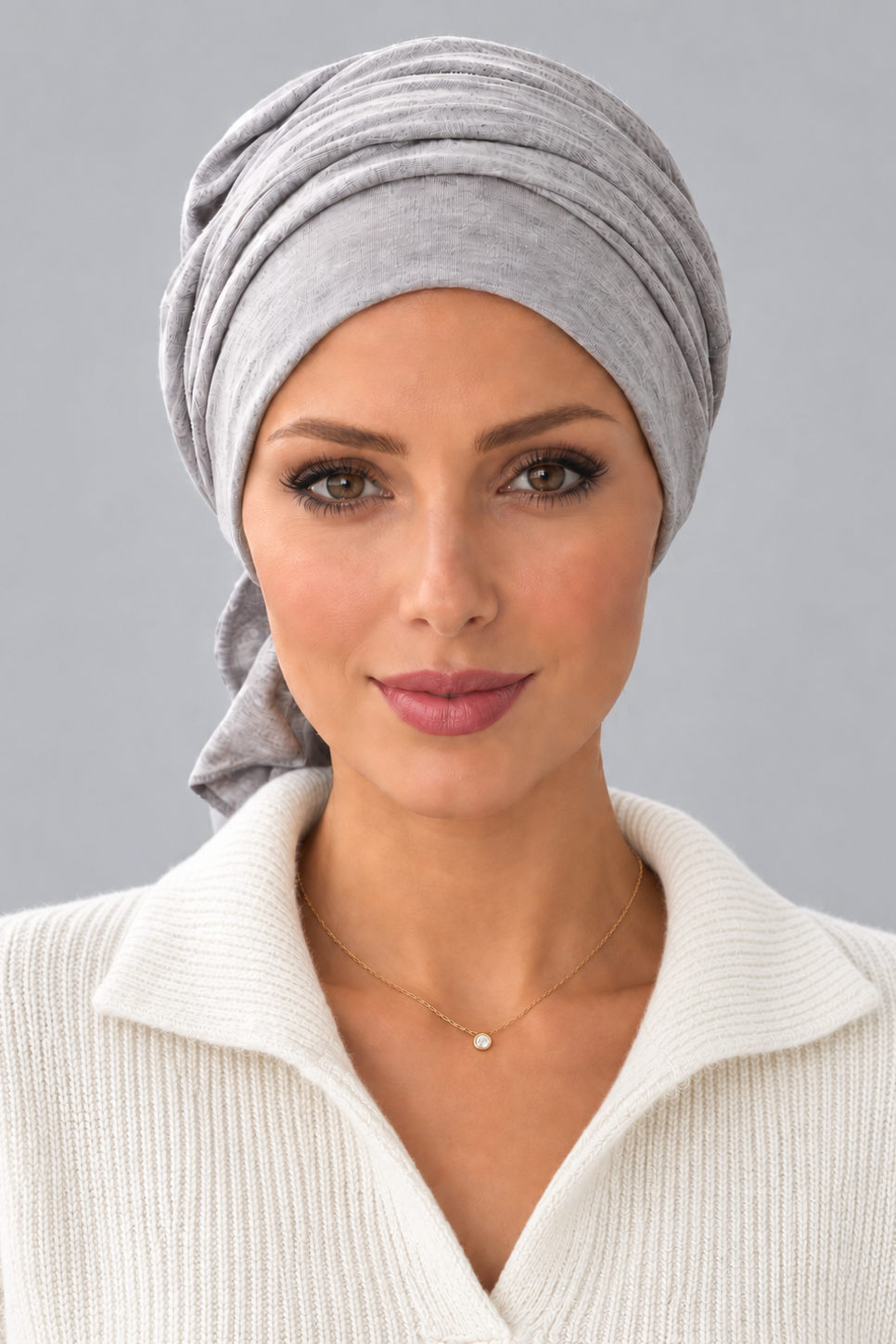 A woman wearing a light gray knitted turban headscarf, paired with a cream ribbed knit top, against a solid gray studio background. Front view