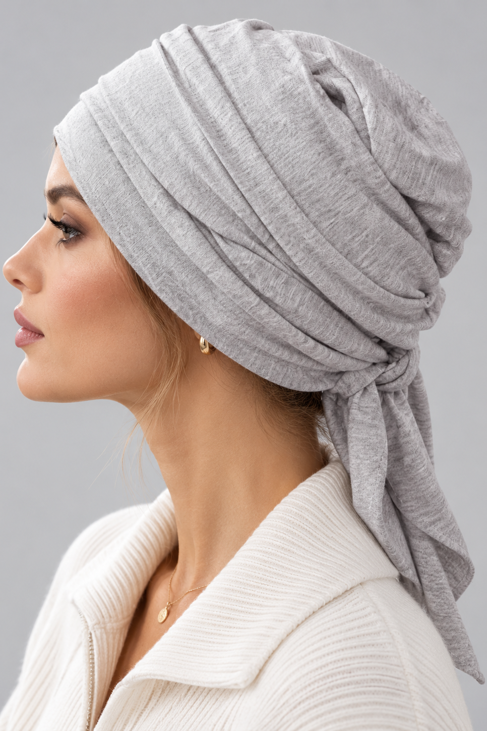 A woman wearing a light gray knitted turban headscarf, paired with a cream ribbed knit top, against a solid gray studio background. Left side view.