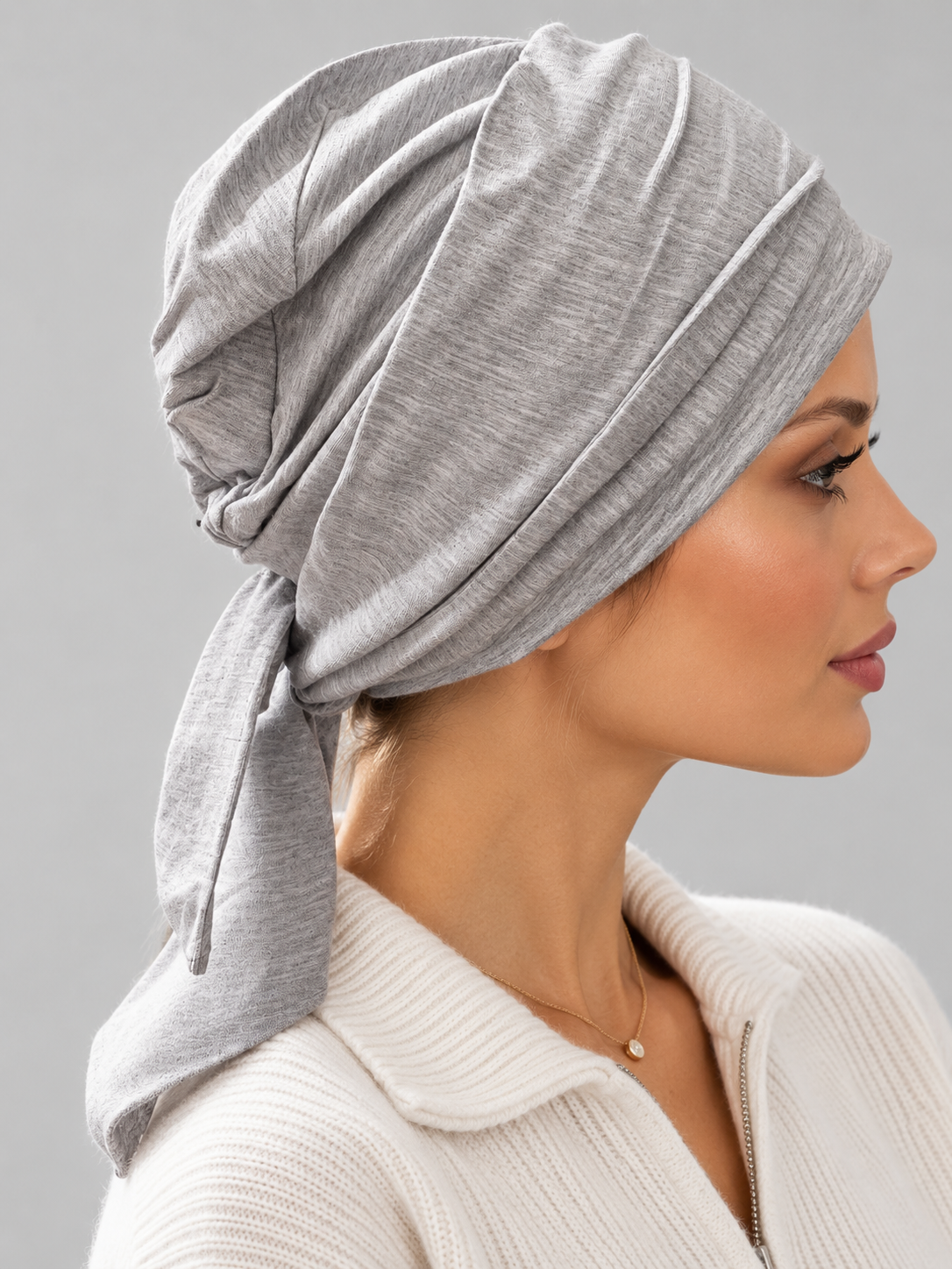 A woman wearing a light gray knitted turban headscarf, paired with a cream ribbed knit top, against a solid gray studio background. Right side view.