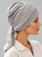 A woman wearing a light gray knitted turban headscarf, paired with a cream ribbed knit top, against a solid gray studio background. Right side view.