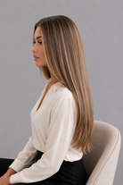Woman sitting in a white chair wearing a Bespoke Human Hair Wig by PUSH in color Jolie and a cream colored button down blouse with black pants. Left side view.