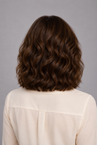 Woman with styled Bespoke Human Hair Wig in a chestnut brown color wearing a white blouse against a gray background. Back view.