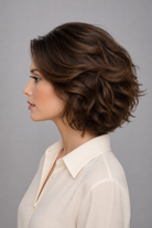 Woman with styled Bespoke Human Hair Wig in a chestnut brown color wearing a white blouse against a gray background. Left side view.