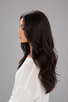 Woman wearing Bradley heat friendly synthetic long wavy wig in color dark chocolate chestnut and a white sweater against a light grey background. Left side view.