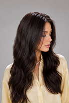 Woman wearing Bradley heat friendly synthetic long wavy wig in color dark chocolate chestnut and a yellow button down blouse against a light grey background. Right side view.