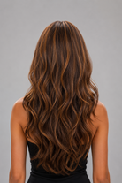 Woman wearing Bradley heat friendly synthetic long wavy wig in color sunlit brunette and a black summer dress with straps against a light grey background. Back view.